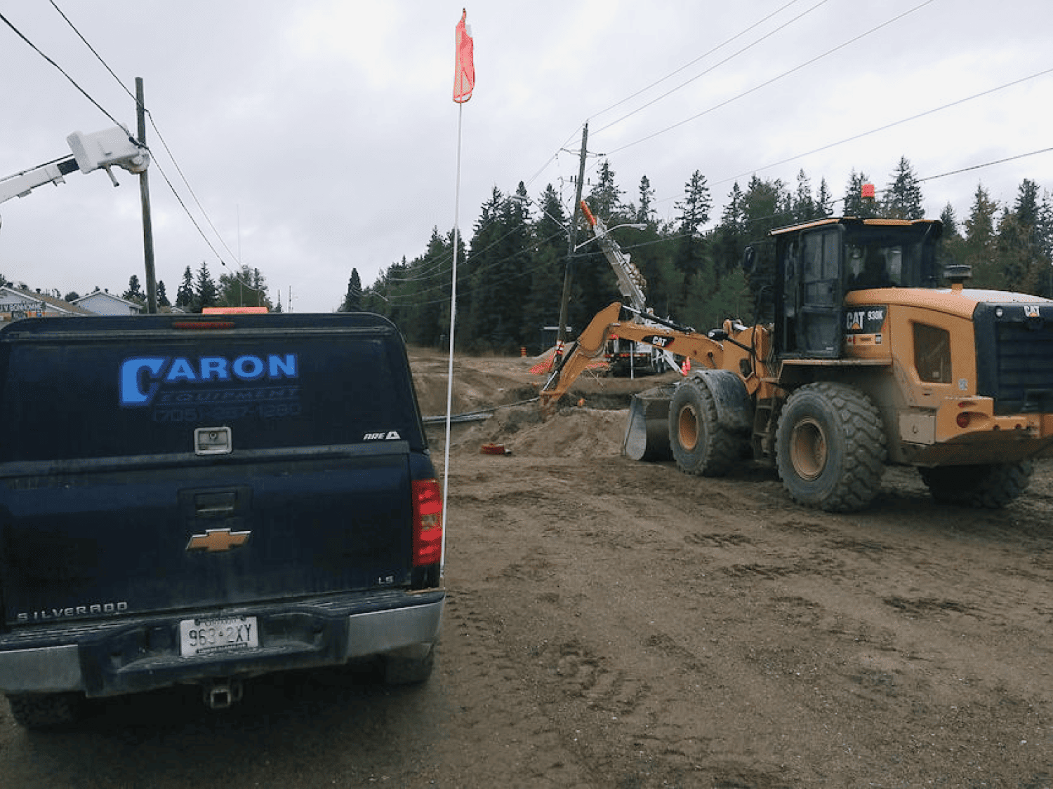 Caron Equipment A bustling scene of civil construction unfolds with Caron Equipment's parked truck and loader on a dirt road. Trees and utility poles stand silhouetted against the cloudy sky, framing the industrious landscape.