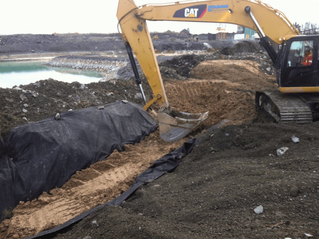 Caron Equipment An excavator from Caron Equipment expertly digs a trench lined with black geotextile fabric at a construction site, enhancing the efficiency of mine site construction with a dirt path and a body of water nearby.