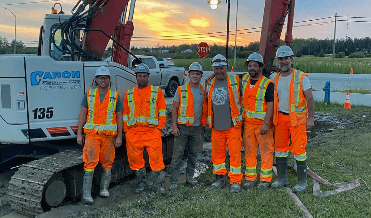 Caron Equipment Six construction workers in orange safety vests and helmets stand in front of an excavator at a roadside construction site during sunset, showcasing expertise akin to mine site construction.