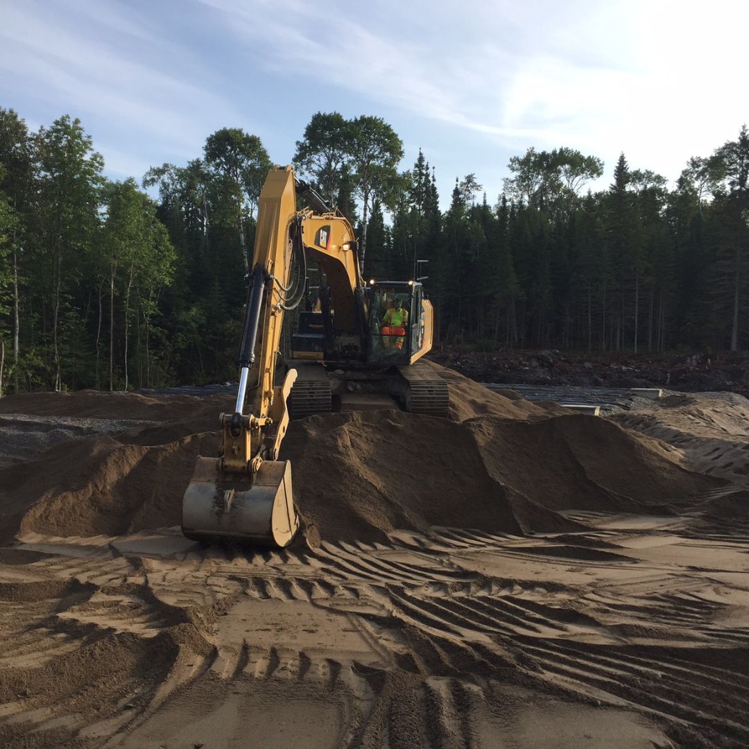 Caron Equipment A yellow excavator from Caron Equipment moves sand on a construction site, surrounded by trees under a cloudy sky.