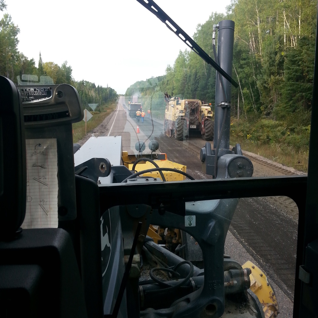 Caron Equipment View from inside a Caron Equipment vehicle cab, showing roadwork with machinery on a forest-lined highway, and cones marking the area.