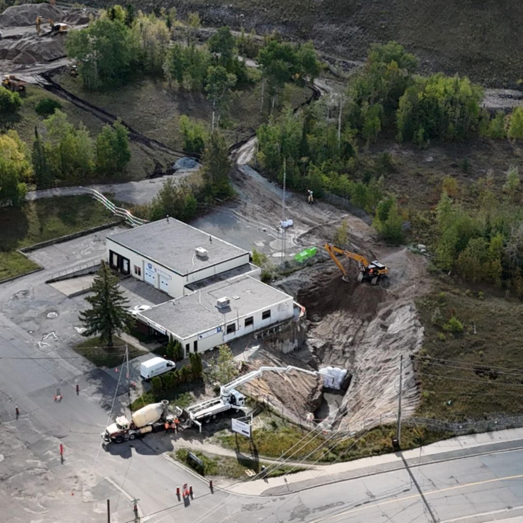 Caron Equipment Aerial view of a construction site with heavy machinery from Caron Equipment around a breached building foundation, surrounded by trees and roads.