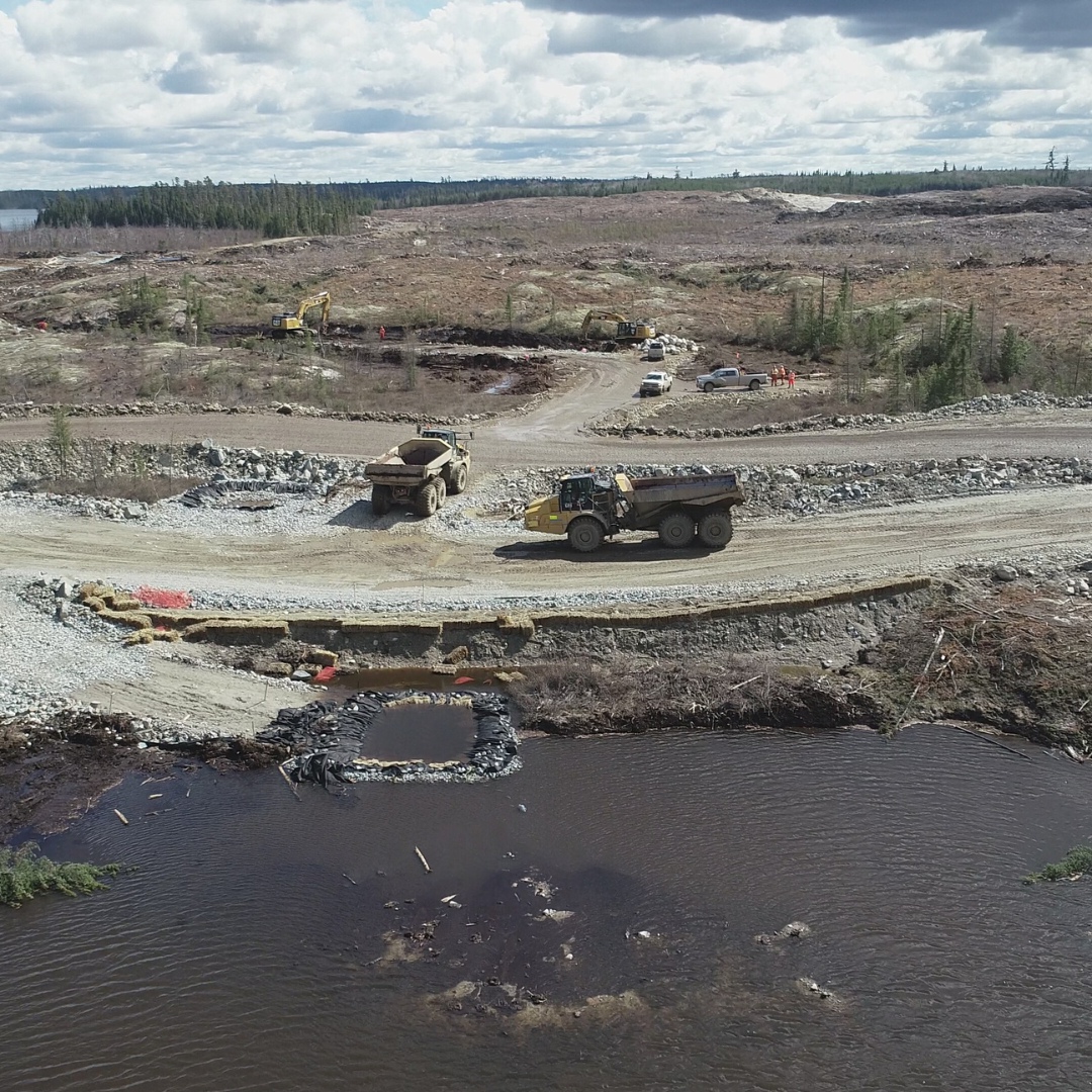 Caron Equipment Aerial view of Civil Construction trucks on a gravel road in a deforested area, with scattered trees and a body of water in the foreground.