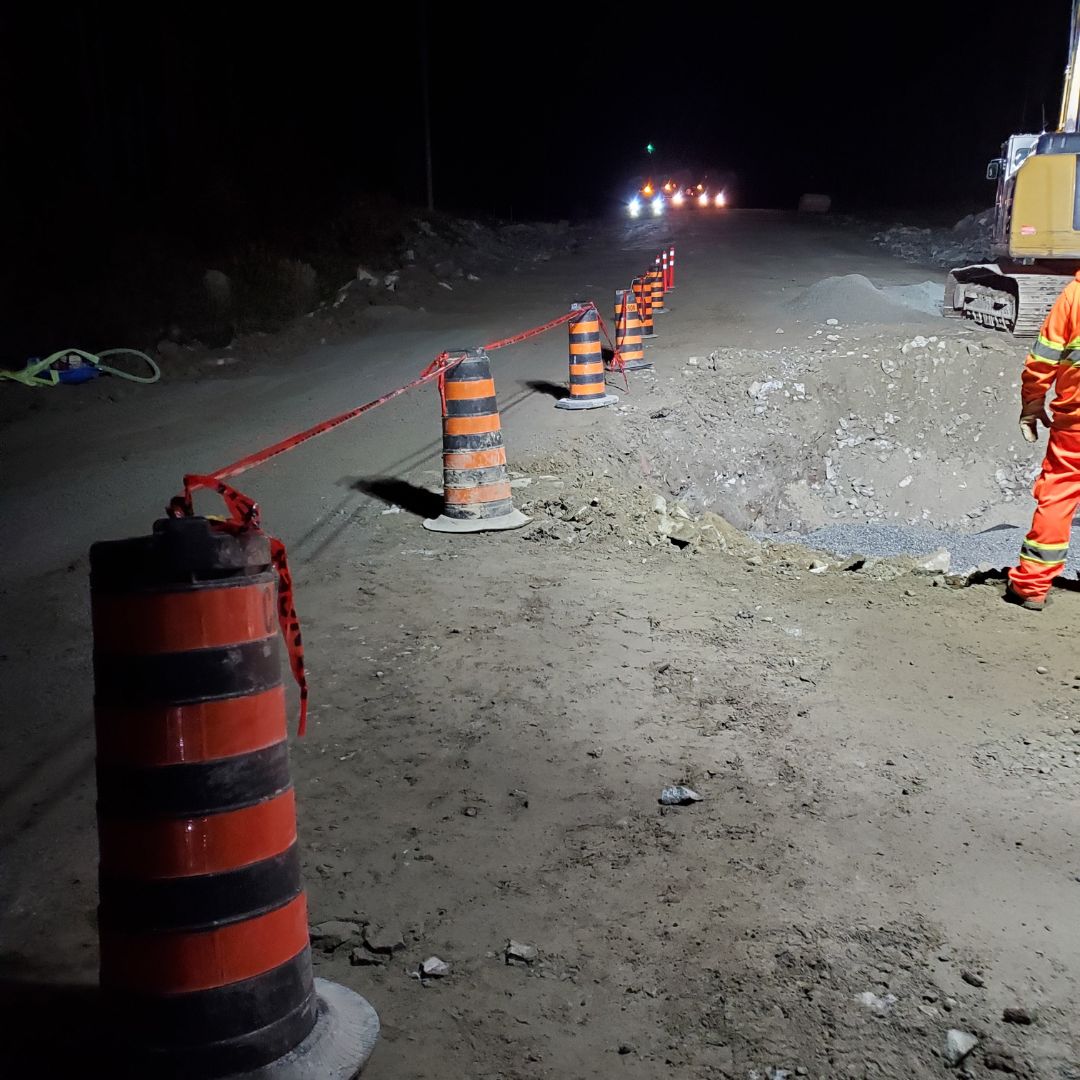 Caron Equipment A construction zone at night with orange cones and red tape marking a large pit. A person in a safety suit stands near an excavator on the dirt road, typifying the precision of mine site construction led by Caron Equipment.