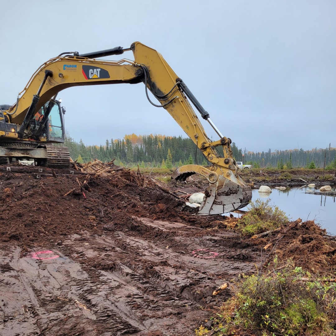 Caron Equipment An excavator from Caron Equipment is steadily digging dirt near a small body of water in a wooded area under a cloudy sky, advancing the progress of an ambitious mine site construction project.