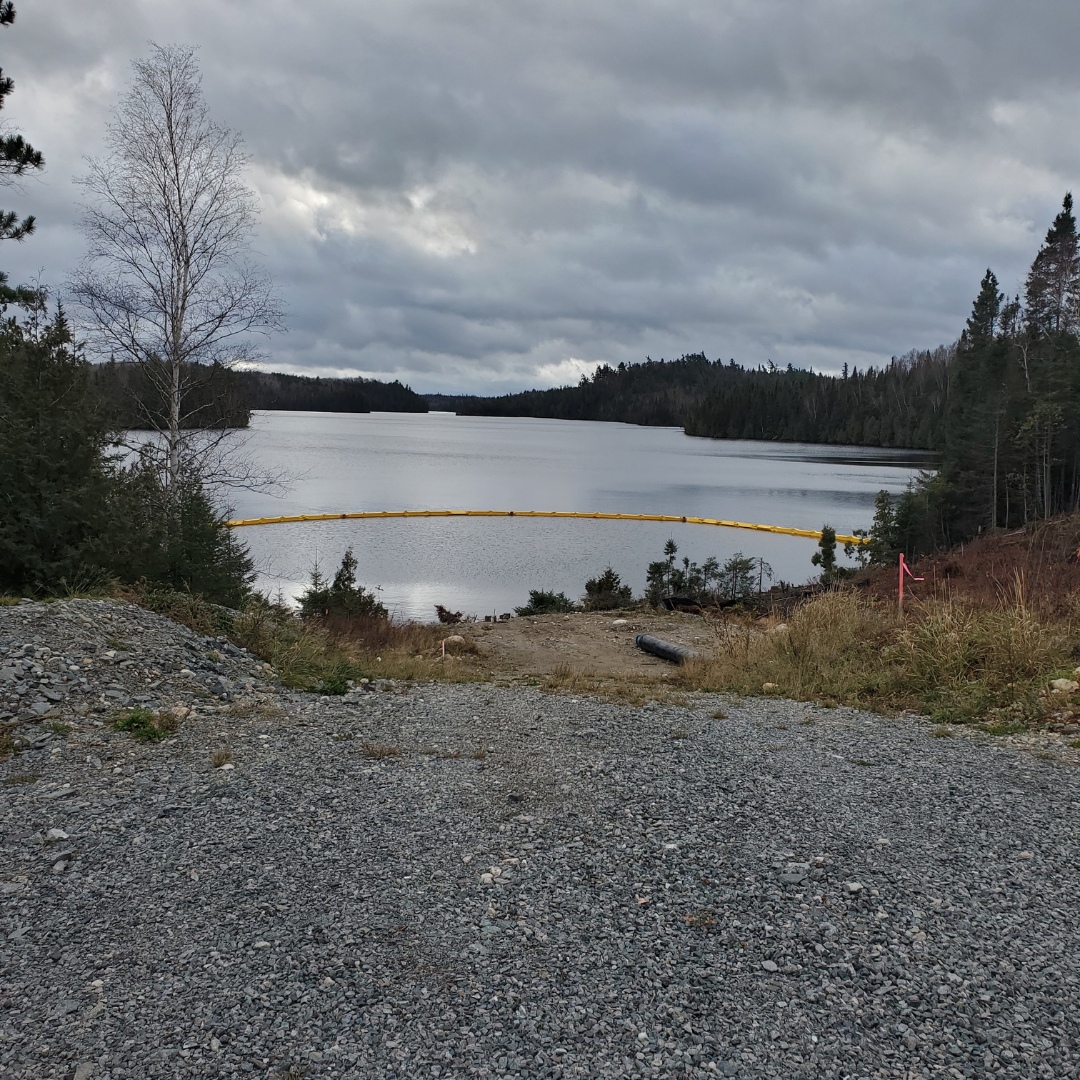 Caron Equipment A gravel path from the former mine site construction leads to a calm lake surrounded by trees under a cloudy sky, with a yellow floating barrier across the water.