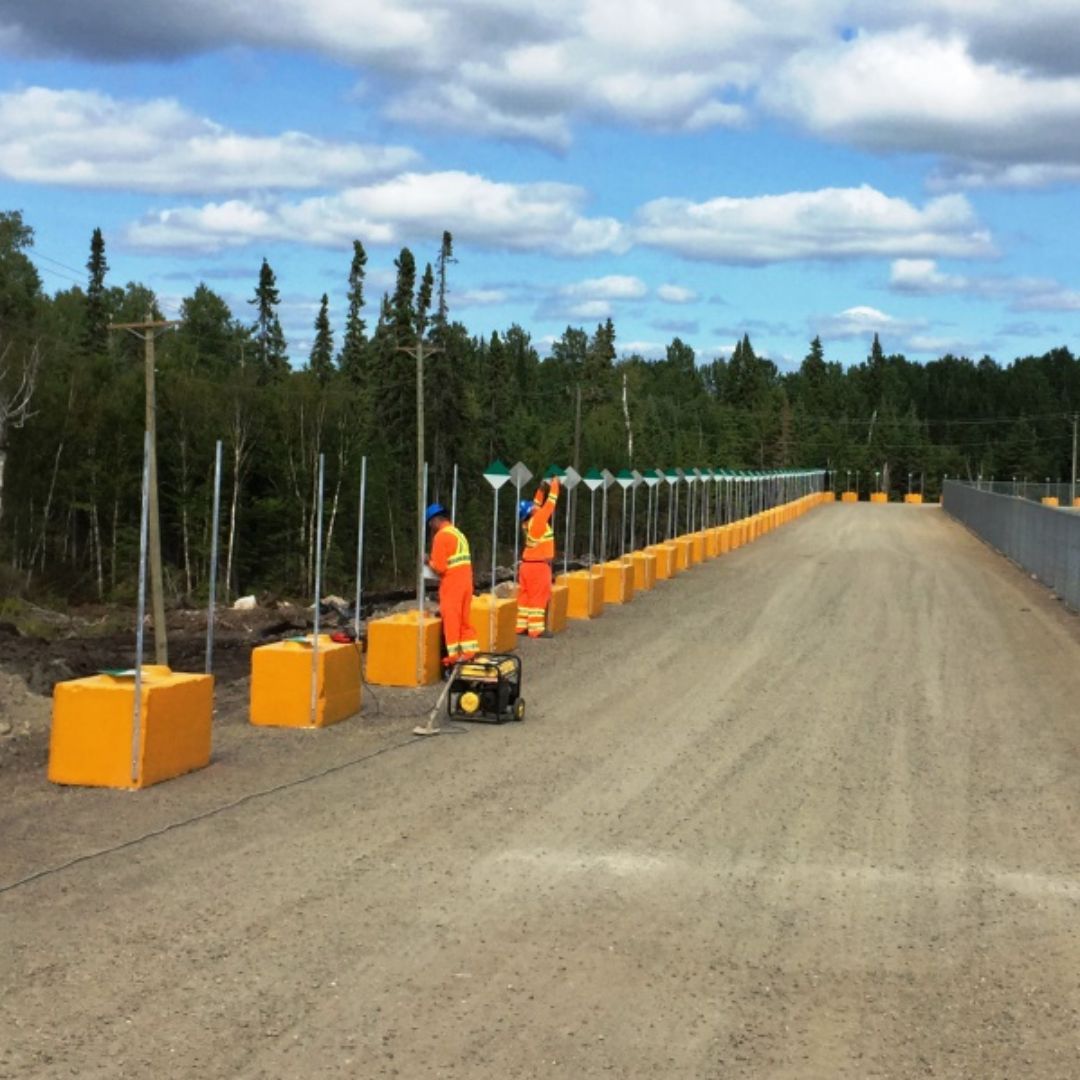 Caron Equipment Two workers in orange uniforms, representing Caron Equipment, install safety barriers along a gravel road surrounded by trees under a partly cloudy sky, showcasing expert Mine Site Construction techniques.