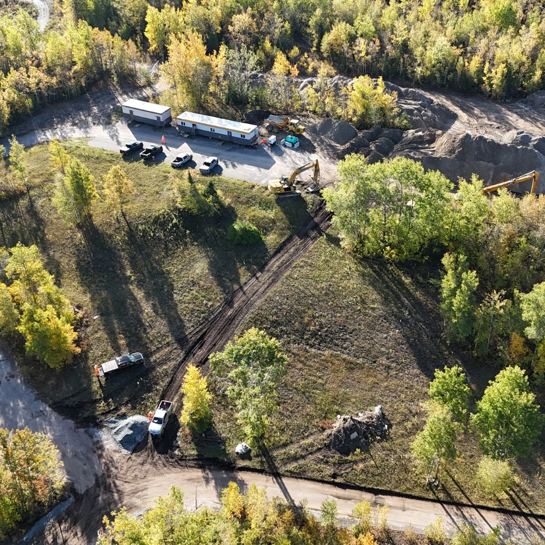 Caron Equipment An aerial view captures a bustling civil construction site with Caron Equipment's machinery, trucks, and trailers nestled amid lush trees and greenery.