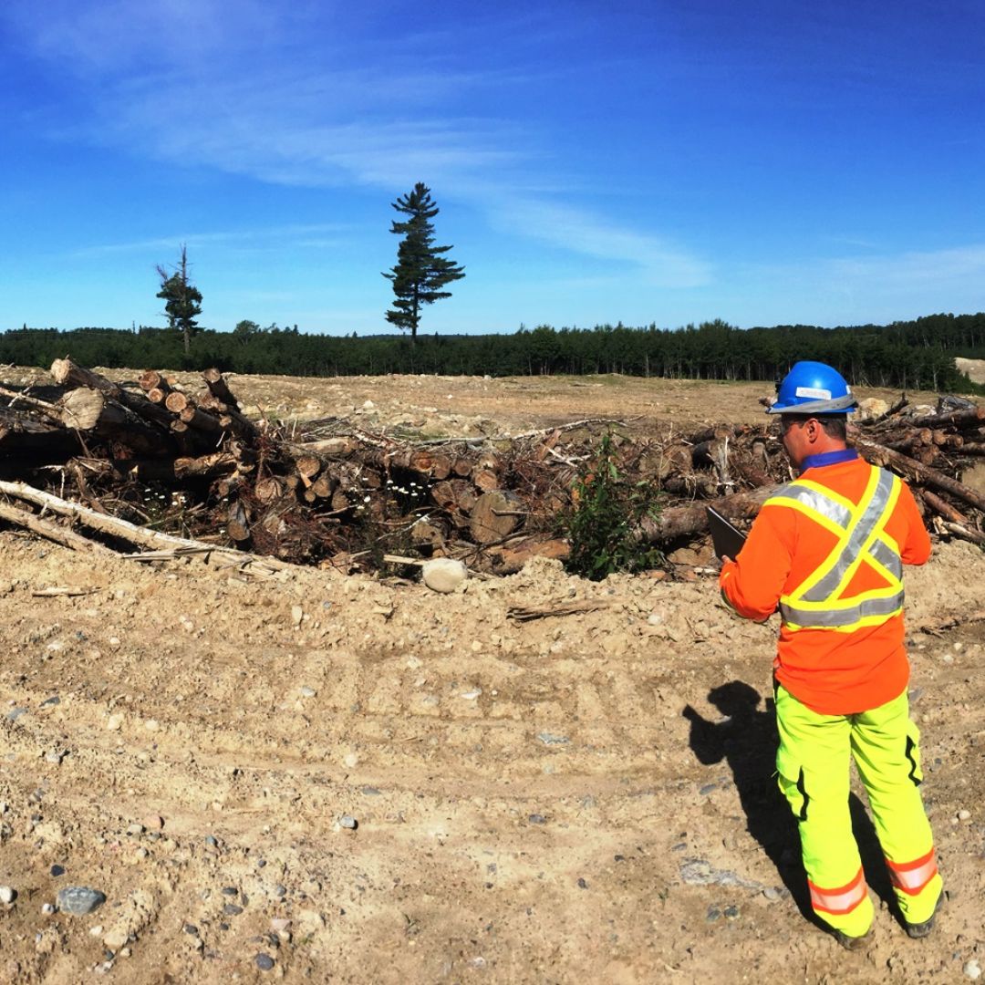 Caron Equipment A worker in high-visibility gear from Caron Equipment examines a civil construction site with piled logs and cleared land under a clear blue sky.