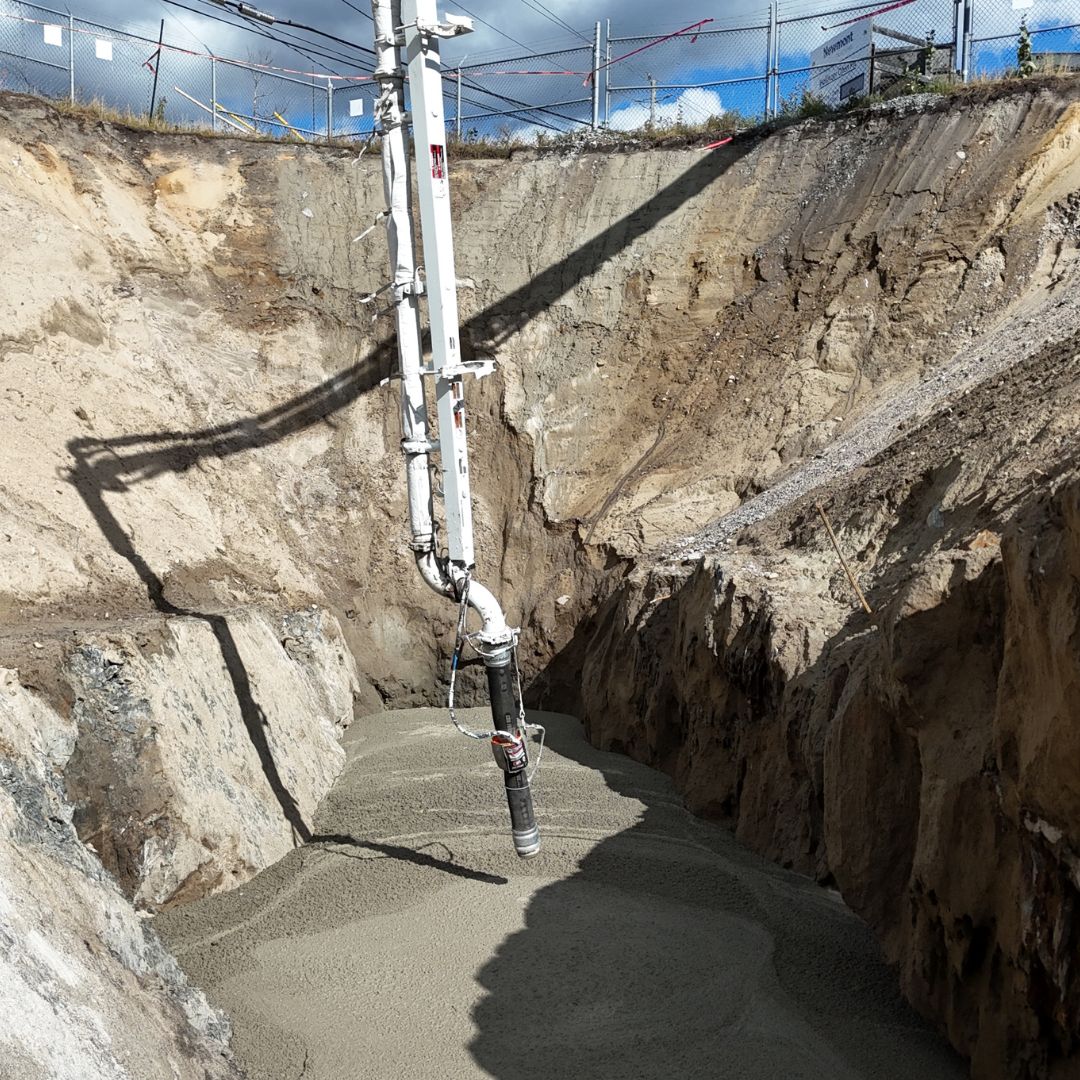 Caron Equipment A large hose pours concrete into a deep construction pit, surrounded by metal fencing. The scene unfolds like a typical day in civil construction, showcasing the efficiency and reliability of Caron Equipment at every step.