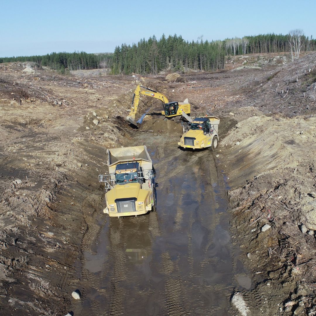 Caron Equipment At the construction site, Caron Equipment’s excavator and two yellow trucks tirelessly navigate the muddy terrain, expertly managing a civil construction project surrounded by cleared land and dense forest.