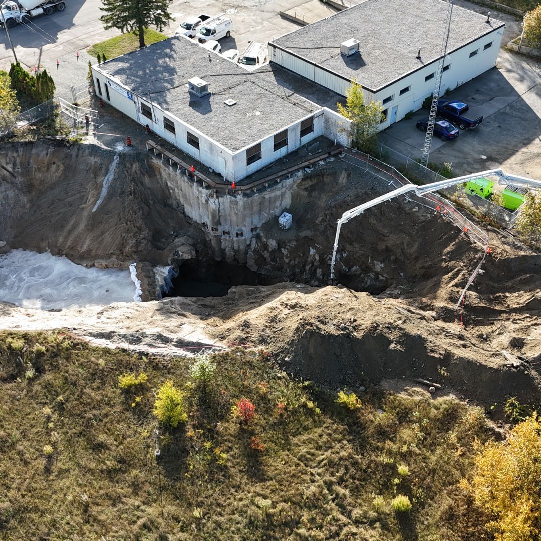 Caron Equipment An aerial view captures the dramatic scene of a large sinkhole near a building, surrounded by construction equipment and vehicles typically seen at a mine site construction area. Caron Equipment machinery is prominently positioned in the adjacent parking zone, ready for civil construction operations.