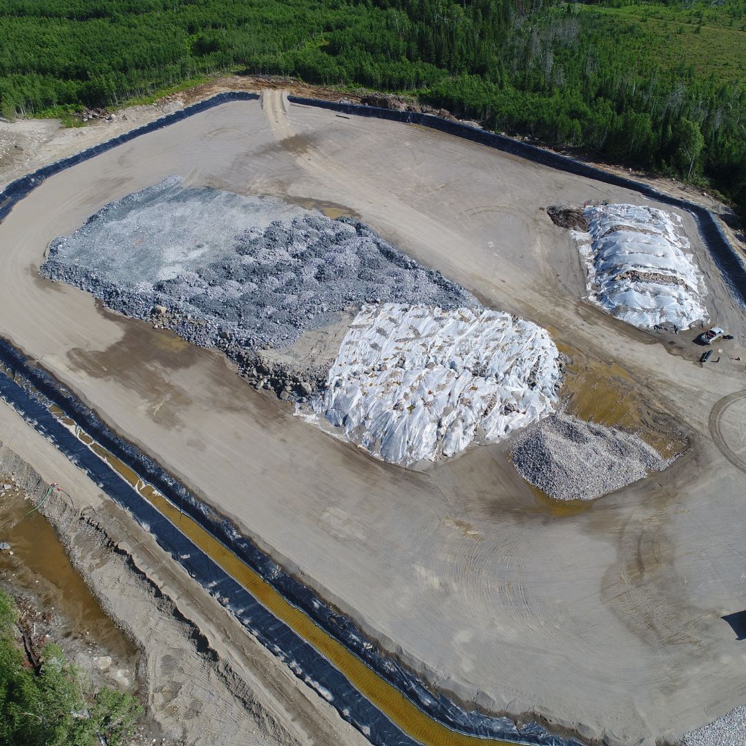 Caron Equipment Aerial view of a large mine site construction area with piles of rocks and covered materials, surrounded by lush green vegetation, showcasing the impressive work of Caron Equipment.