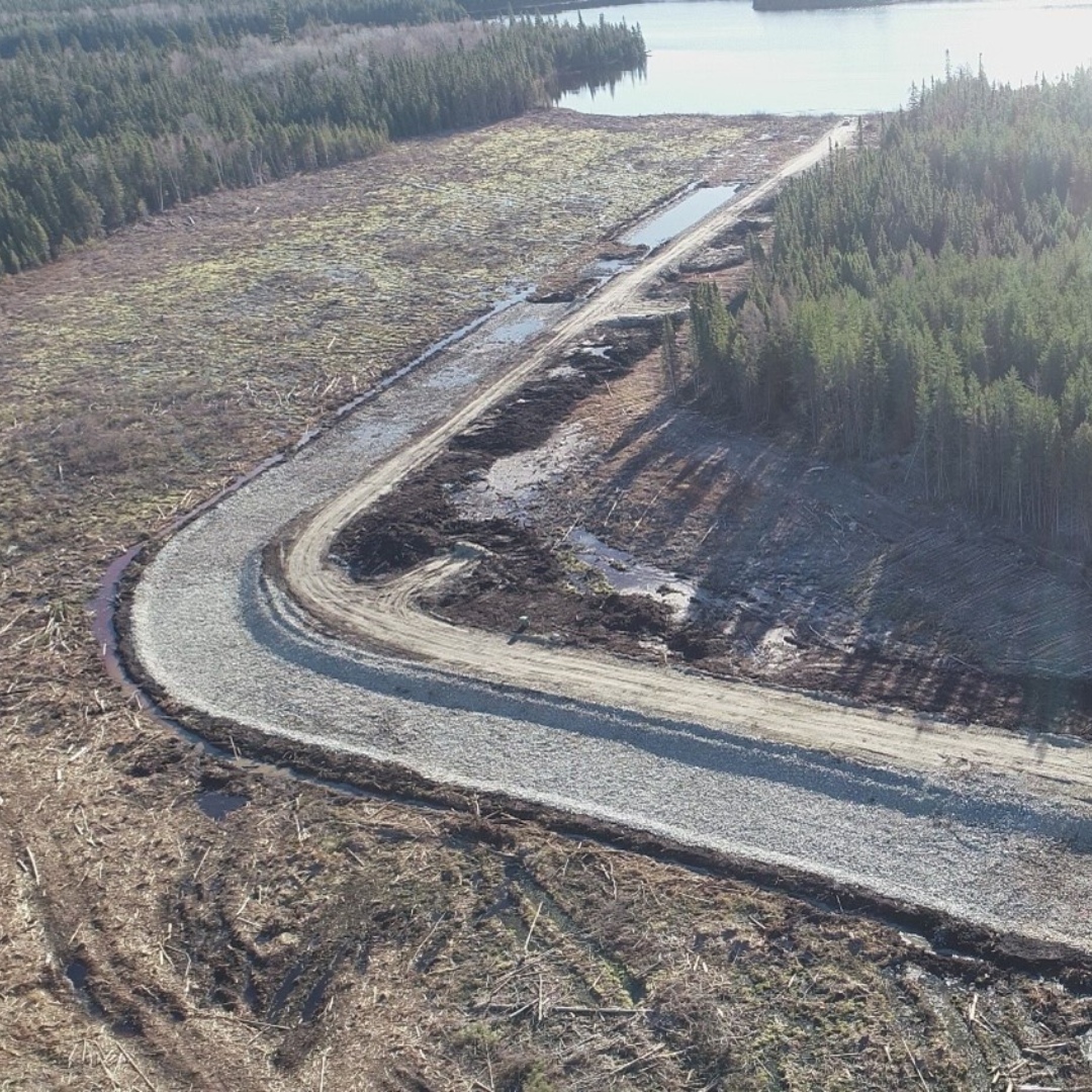 Caron Equipment An aerial view reveals a gravel road winding through a deforested expanse near a lake, framed by coniferous trees—a testament to Caron Equipment's expertise in civil construction.