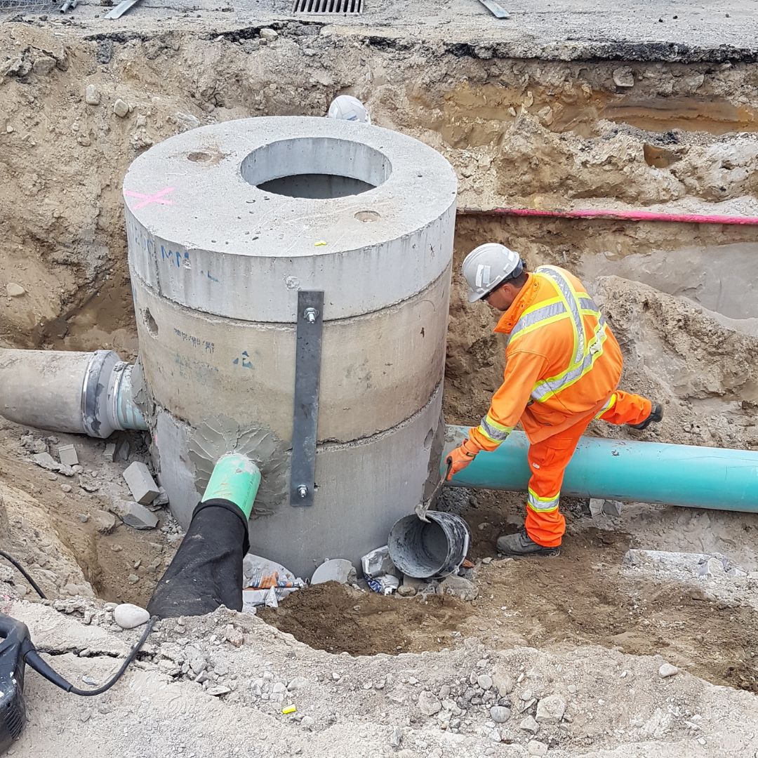 Caron Equipment A construction worker in orange safety gear inspects a concrete drainage structure with connecting pipes at a civil construction site, featuring Caron Equipment's latest innovations.