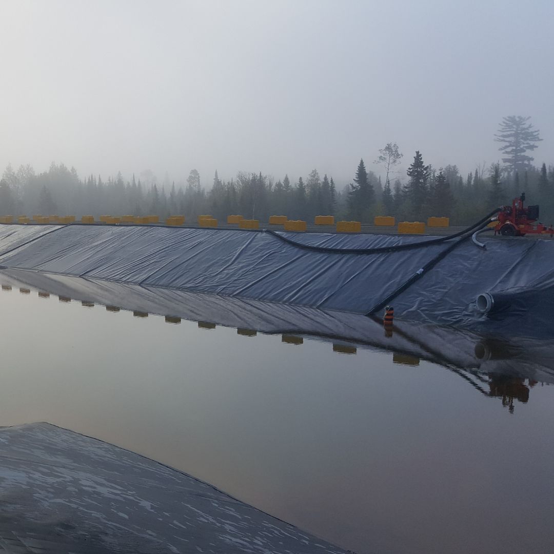 Caron Equipment A large plastic-covered embankment with machinery and hoses at the top marks the Caron Equipment mine site construction, surrounded by reflective water and a misty forest background.