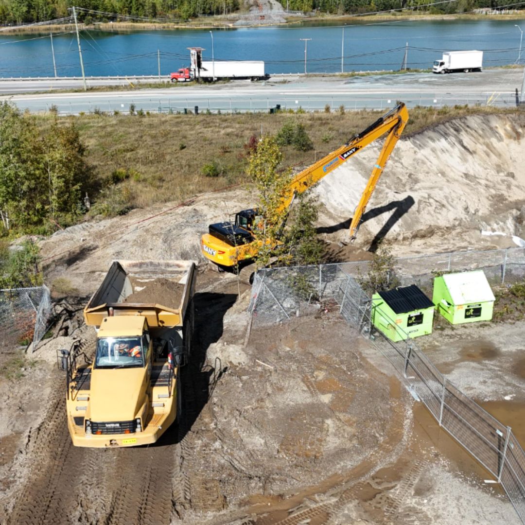 Caron Equipment At a bustling mine site construction area, a massive excavator from Caron Equipment loads dirt into a dump truck, while nearby vehicles pass by on the road and a scenic river winds through the background.