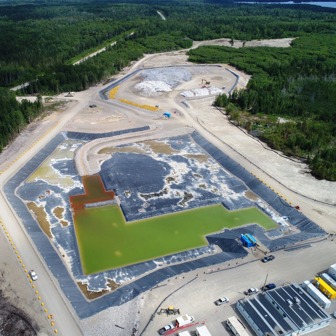 Caron Equipment Aerial view of a mine site construction with a large green water-filled area lined with black tarp and surrounding machinery, set amidst a forested landscape.