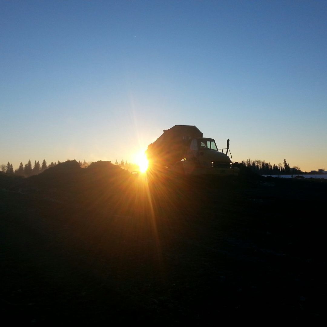 Caron Equipment Silhouette of a dump truck from Caron Equipment against a setting sun, with mounds of earth typical of mine site construction and a clear sky in the background.