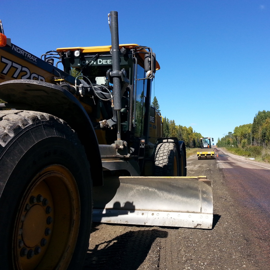 Caron Equipment A road grader diligently works on a rural path, flanked by trees under a clear blue sky. In the distance, another machine stands ready, echoing the meticulous precision often seen in civil construction projects by Caron Equipment.