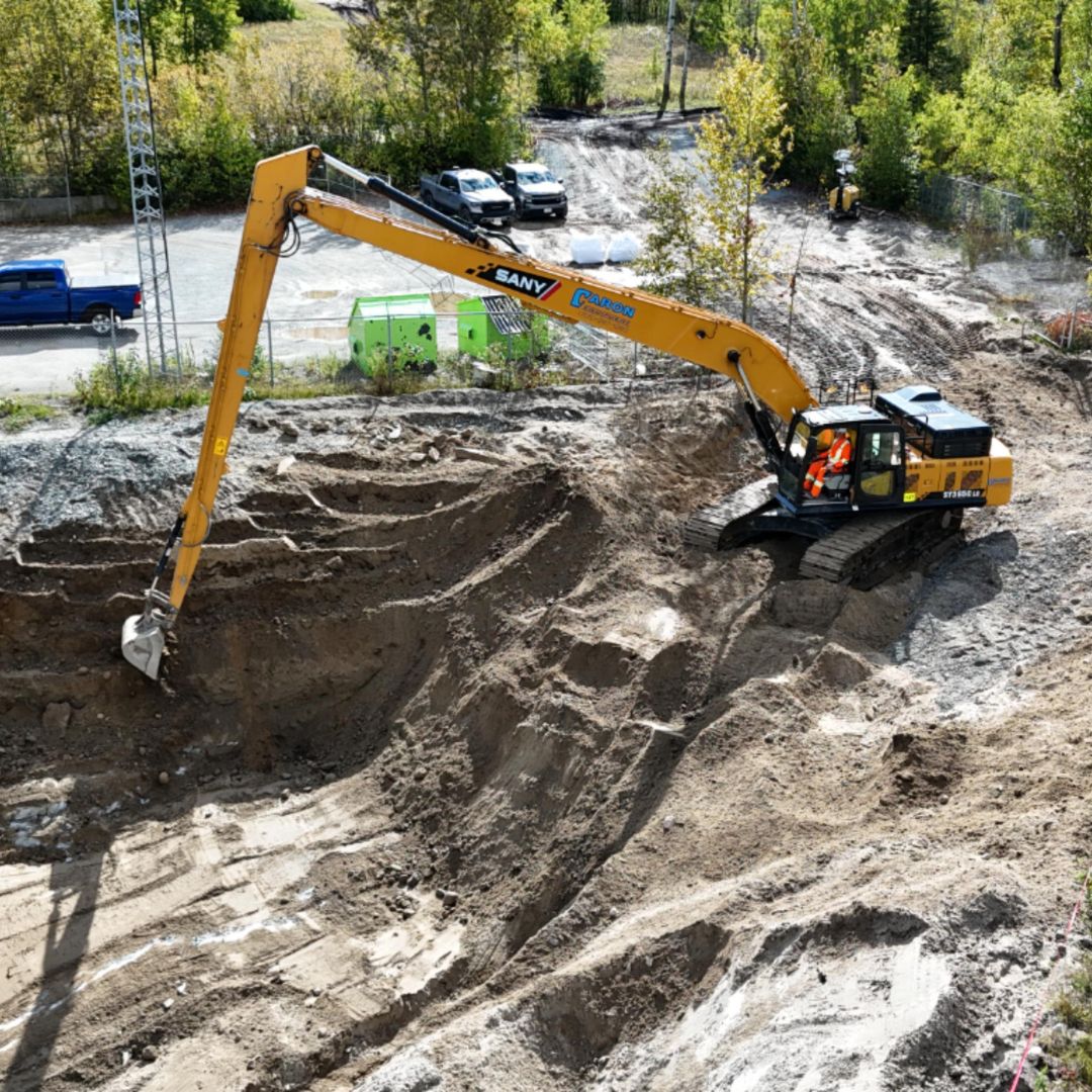 Caron Equipment A yellow excavator with a long arm digs diligently at a sprawling mine site construction. Inside the cab, two workers in orange vests collaborate efficiently, surrounded by vehicles and trees in the background—a testament to Caron Equipment's expertise in civil construction.