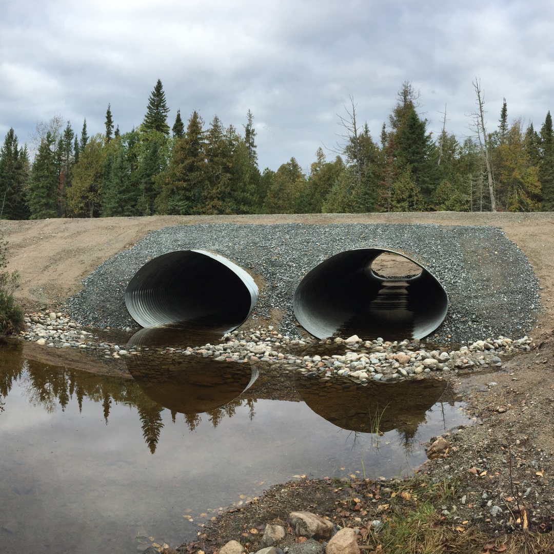 Caron Equipment Two large metal culverts with a rocky base facilitate water flow under a gravel road in a forested area. This civil construction project by Caron Equipment ensures durability and efficient drainage, all beneath the cloudy sky above.