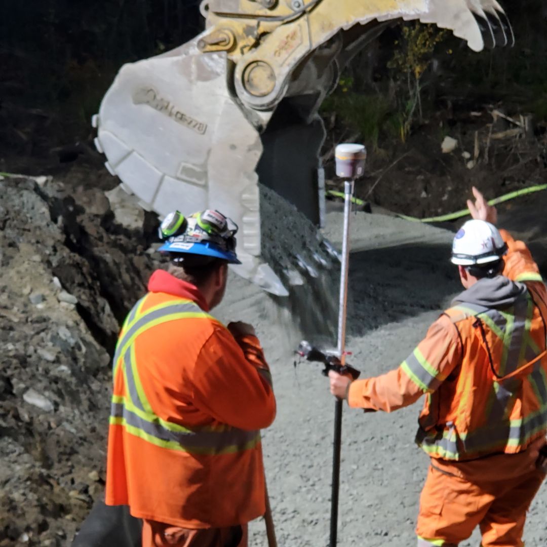 Caron Equipment Two construction workers in safety gear, from Caron Equipment, direct a large excavator pouring gravel onto a road. One worker holds a measuring rod, showcasing expertise in civil construction.