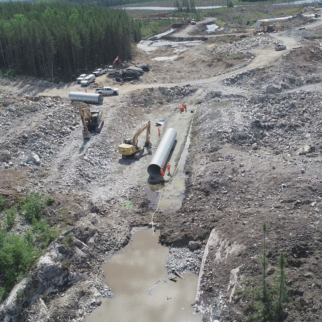 Caron Equipment At a robust construction site, Caron Equipment's machinery diligently installs a large pipeline in the rocky, forested terrain. Workers in orange vests coordinate operations as a muddy river meanders through this bustling scene of civil construction expertise.