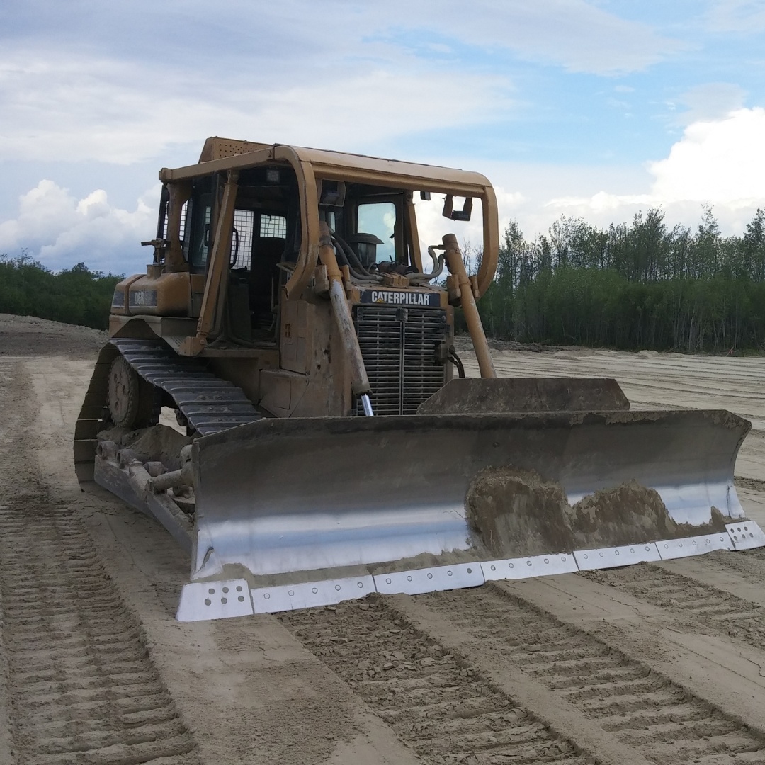 Caron Equipment A bulldozer from Caron Equipment sits on a dirt construction site, its blade lowered and ready for action. The scene is framed by trees and a cloudy sky, capturing the essence of civil construction in harmony with nature's backdrop.