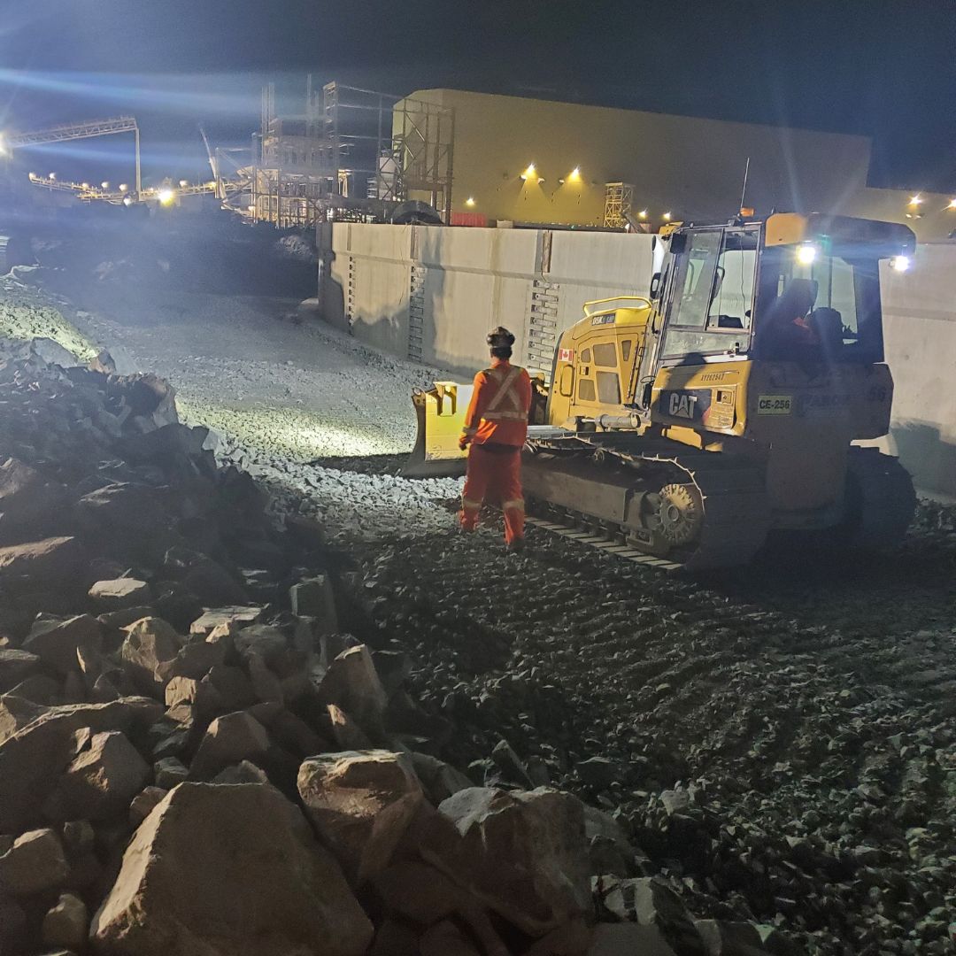 Caron Equipment A construction worker in high-visibility clothing stands near heavy machinery at a bustling mine site construction, with rocks and a lit industrial landscape in the background at night.