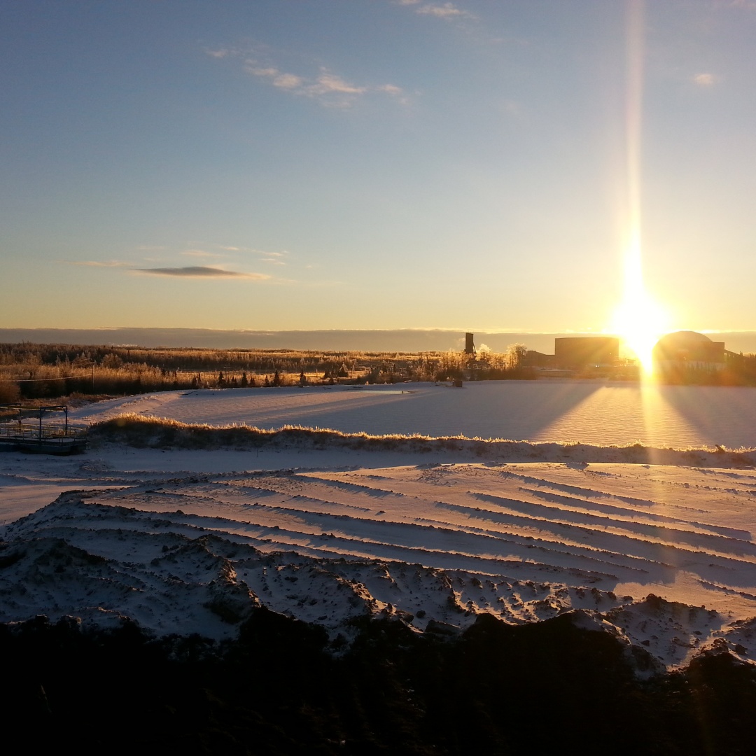 Caron Equipment At sunrise, the snowy field stretches under a clear sky with the sun shining brightly over the horizon. Silhouettes of buildings and Caron Equipment hint at nearby civil construction work, crafting a serene yet industrious landscape.