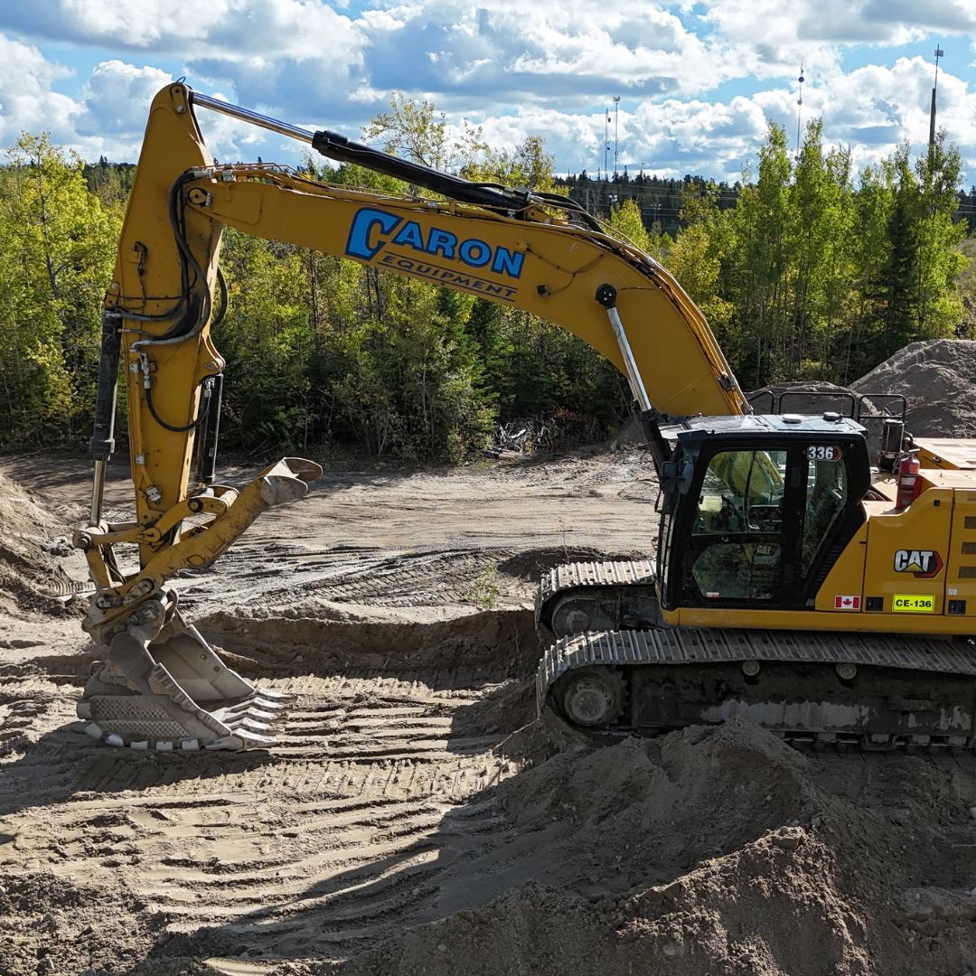 Caron Equipment A yellow excavator from Caron Equipment is diligently digging in a sandy civil construction site surrounded by trees under a partly cloudy sky.