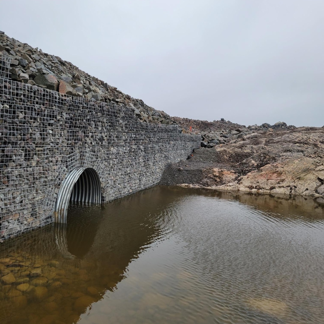 Caron Equipment A concrete culvert, a testament to civil construction, drains into a pool of water surrounded by rocky terrain under an overcast sky.