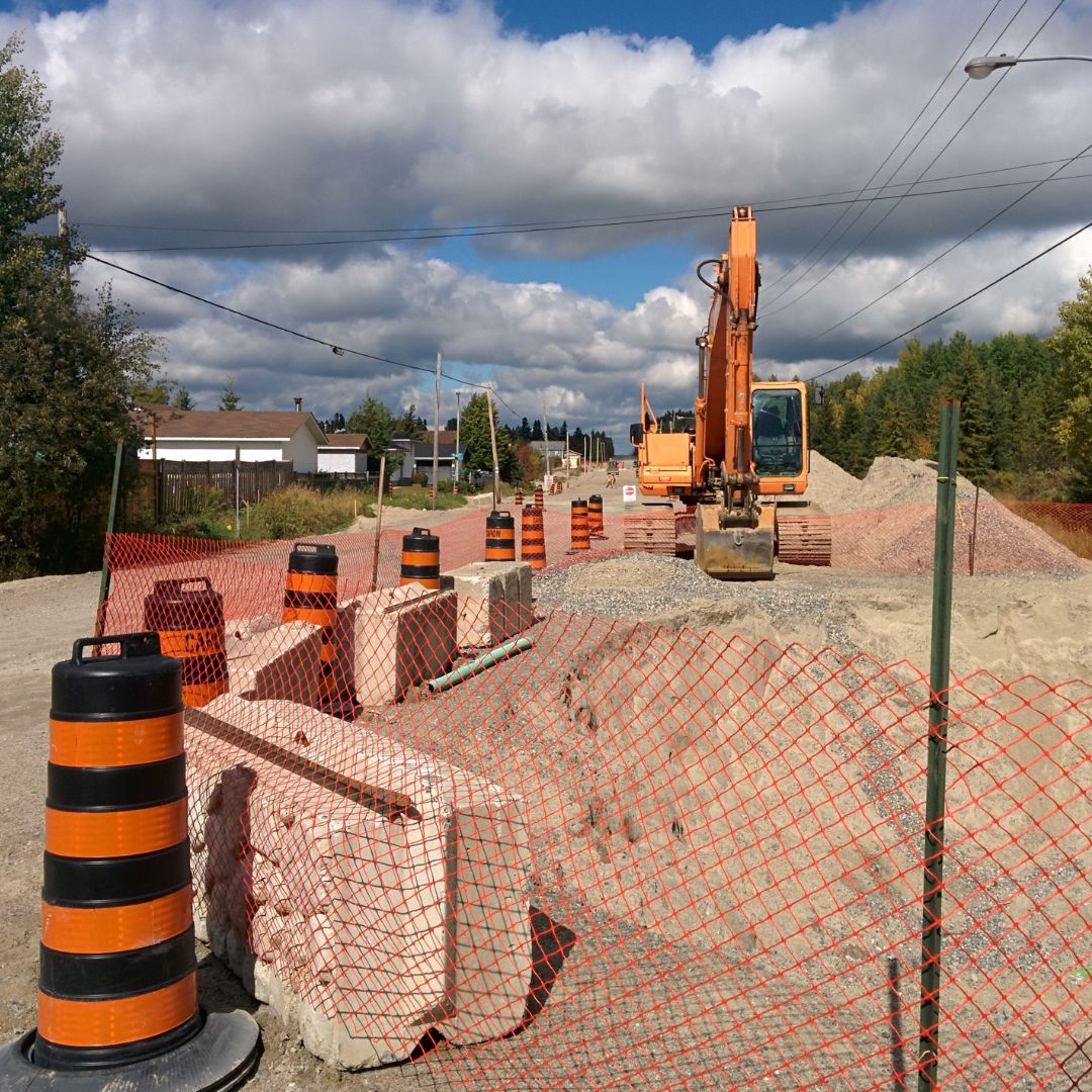 Caron Equipment The construction site, reminiscent of civil construction projects, features an excavator on a gravel road surrounded by orange barrels and a safety net, set under a partly cloudy sky. With precision akin to Caron Equipment's standards, the scene reflects an organized blend of industry and nature.