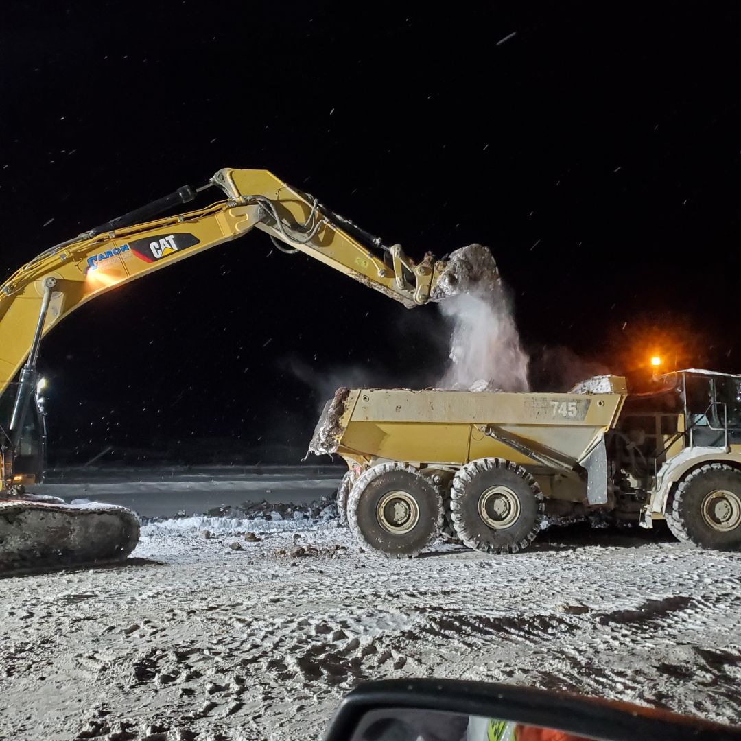 Caron Equipment A large Caron Equipment excavator loads dirt into a dump truck at a snowy civil construction site at night.