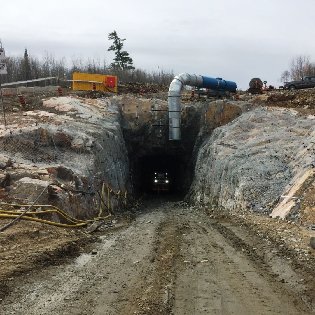 Caron Equipment Entrance to a rock tunnel with Caron Equipment parked beside it and a vehicle inside. The civil construction area has a dirt path leading to the tunnel under an overcast sky.