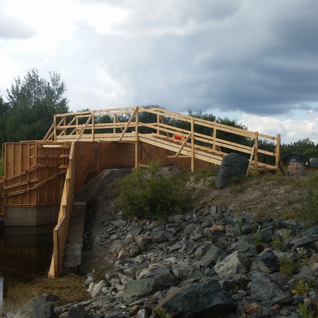 Caron Equipment A wooden footbridge, part of a civil construction project by Caron Equipment, is under construction. It spans a rocky bank beneath cloudy skies in the background.