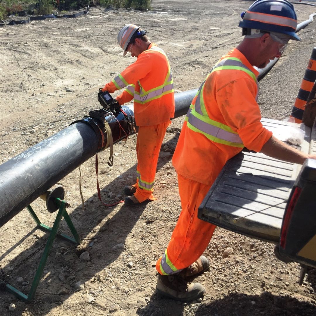 Caron Equipment Two workers in orange safety gear inspect a pipeline in a civil construction area. One checks equipment from Caron Equipment while the other reads documents on a truck tailgate.