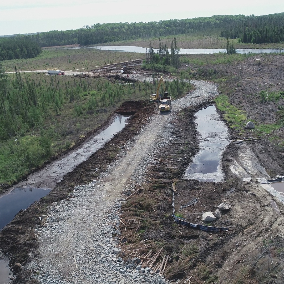 Caron Equipment Caron Equipment's machinery stands on a partially built road winding through a forested area, with cleared land and water flanking either side.