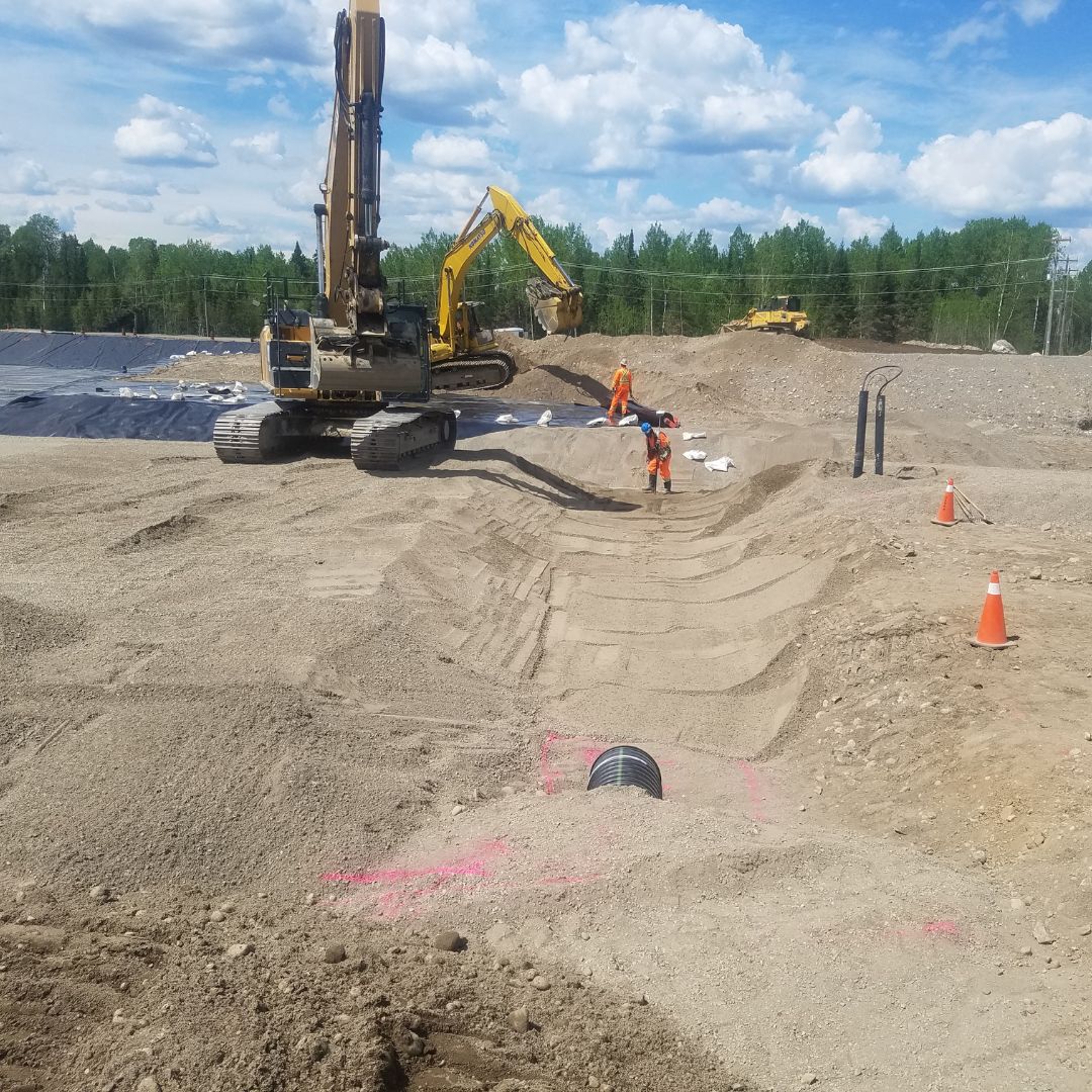 Caron Equipment On a civil construction site, excavators and workers in orange safety gear navigate the sandy terrain. Caron Equipment marks the area with cones while a pipe is visible in the foreground under the cloudy sky overhead.