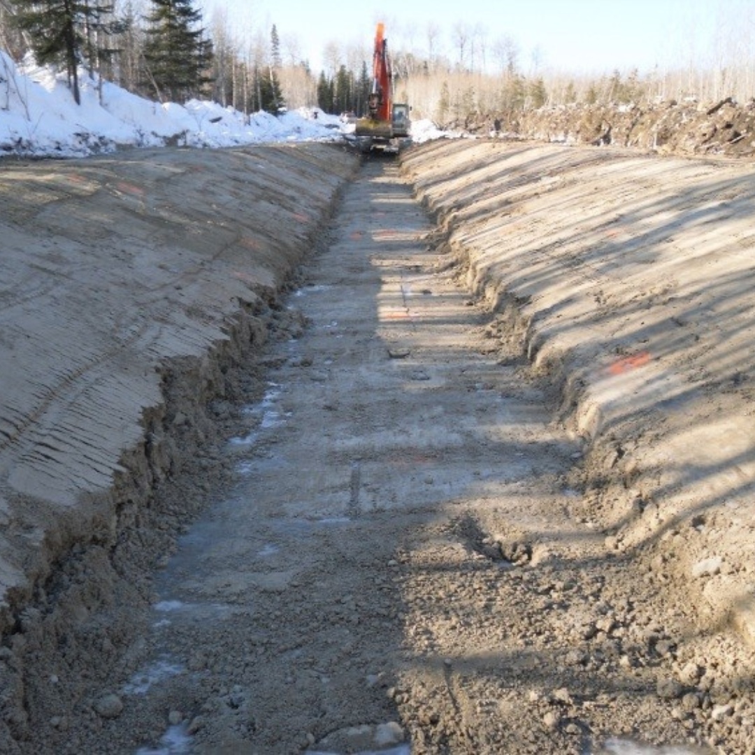 Caron Equipment An excavator, branded with Caron Equipment, is expertly digging a trench in a snowy, forested area. The trench, part of a civil construction project, is lined with snow and surrounded by towering trees.