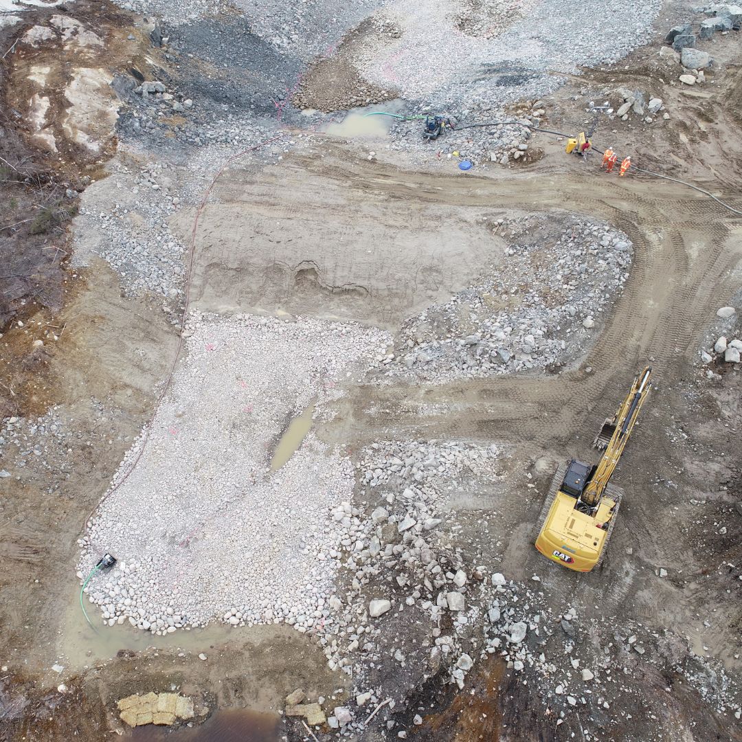 Caron Equipment Aerial view of a civil construction site with a large excavator and four workers in safety gear. The ground is covered with rocks and dirt, typical of mine site construction projects managed by Caron Equipment.