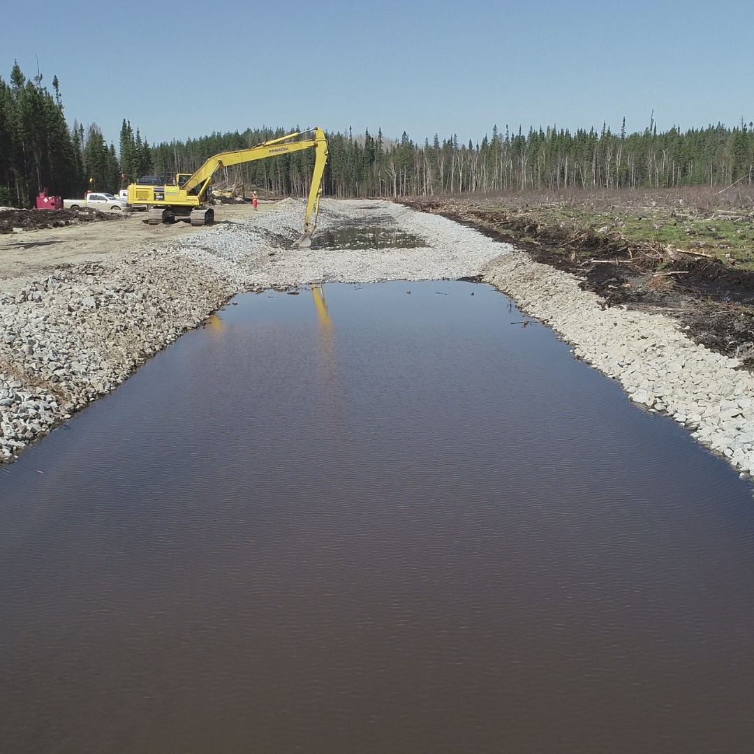 Caron Equipment An excavator from Caron Equipment is diligently working near a newly constructed canal lined with rocks. Surrounded by a dense forest and construction equipment, this civil construction project exemplifies excellence in mine site development.