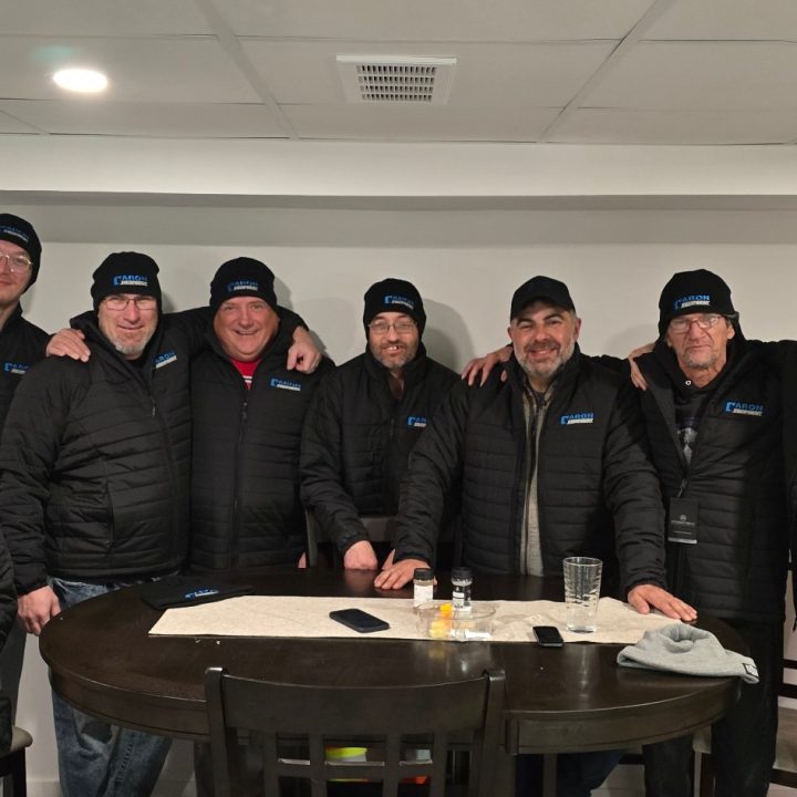 Caron Equipment A group of ten people wearing matching jackets and hats stand around a table indoors, discussing plans for the upcoming mine site construction project.