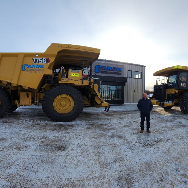 Caron Equipment Two large yellow dump trucks, branded with Caron Equipment, are parked on snowy ground in front of a building. A person stands between the trucks, emphasizing their role in civil construction activities.