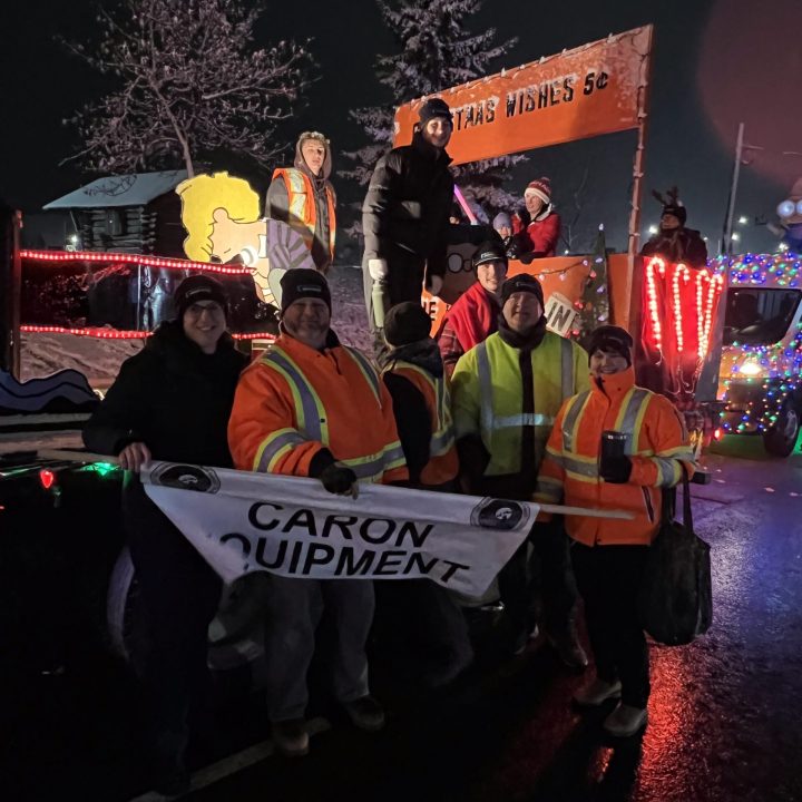 Caron Equipment A group of people in reflective jackets stand and sit on a lit-up parade float adorned with Christmas lights, proudly holding a "Caron Equipment" banner. This cheerful display highlights Caron Equipment's expertise in mine site construction under the night sky.