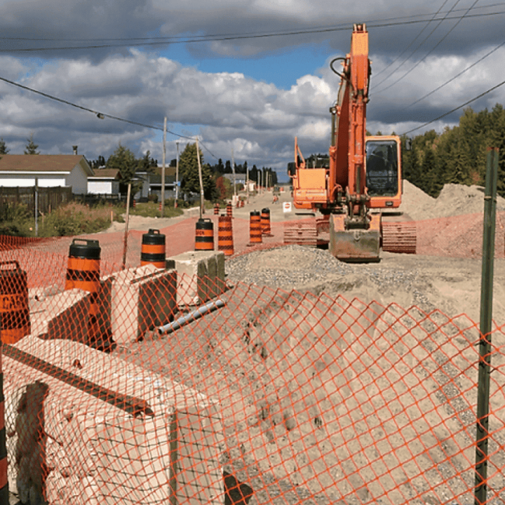 Caron Equipment A construction site on a dirt road features an excavator, traffic barrels, and orange fencing—hallmarks of a Caron Equipment project. Piles of gravel are visible under the partly cloudy sky, showcasing their expertise in civil construction.